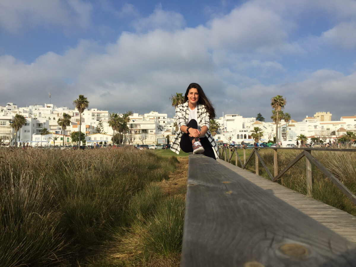 Irene sentada en barandilla camino de la playa con le pueblo de fondo